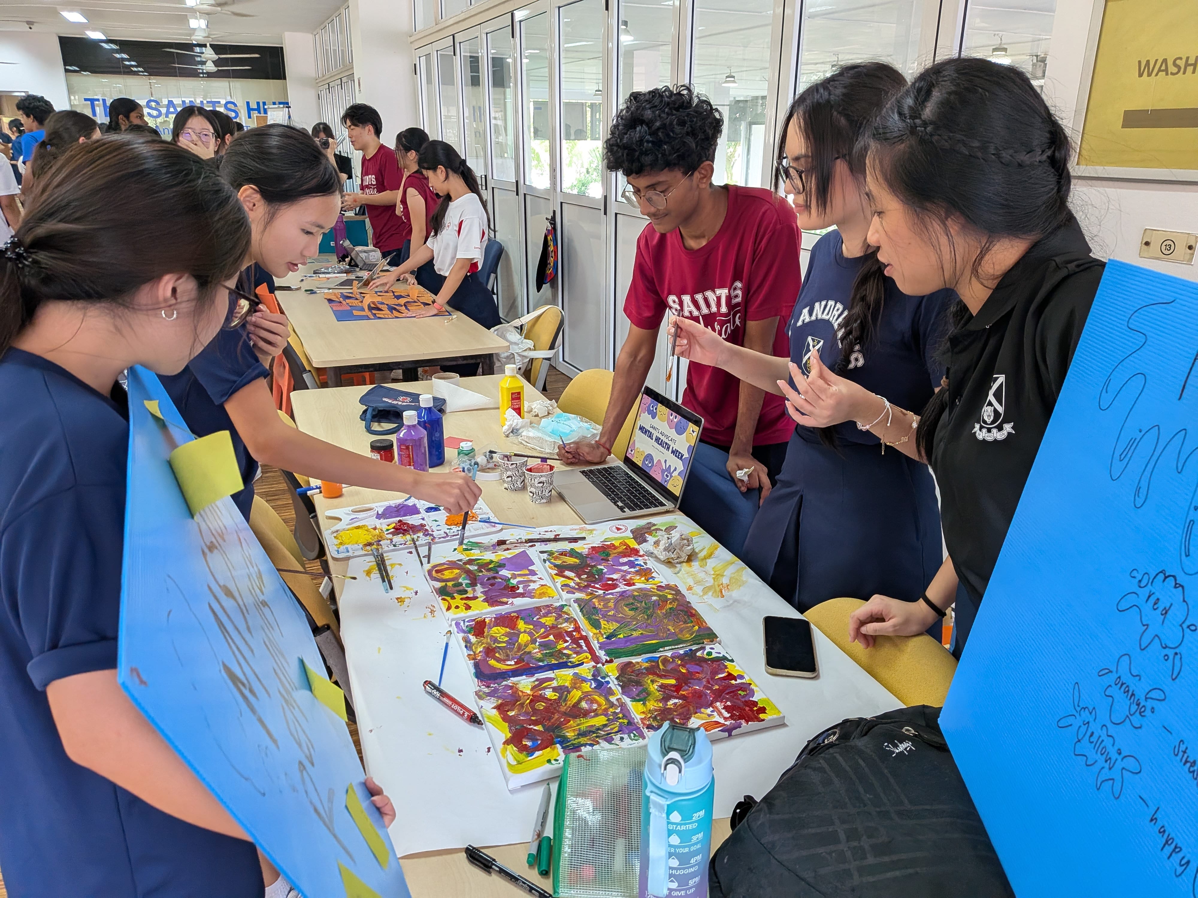 Students collaborating around a table filled with colorful painted canvases during a Mental Health Week art booth activity.